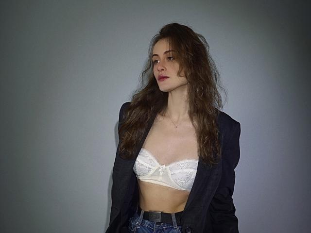 AlesyaDrons with long brown hair, wearing a black blazer over a white lace bra, and blue jeans, standing against a gray wall, showing her small tits.