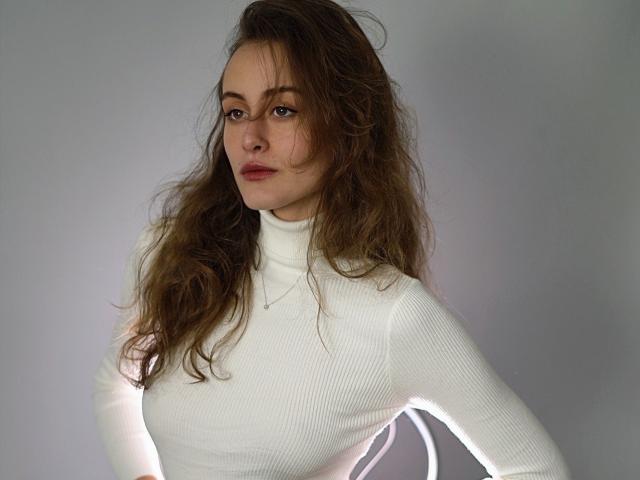 Alesya Drons hat welliges, braunes Haar, ein weißes Rollkragenoberteil und einen ernsten Gesichtsausdruck. Ihre Brüste sind unter dem engen Oberteil sichtbar, und sie trägt eine zarte Halskette..