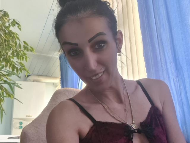 SexyAnnaNatural mit dunklem Haar zu einem Dutt gebunden, dicken Augenbrauen, Nasenring, lächelnd, trägt einen roten Spitzen-BH, eine Halskette, im Hintergrund sind blaue Vorhänge und eine Topfpflanze zu sehen..