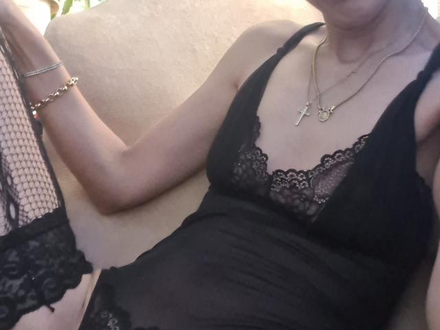 SexyAnnaNatural, con pechos pequeños, vistiendo lencería de encaje negro y un vestido negro transparente, sentada en un sofá, con un collar dorado con una cruz, una pulsera y una manga de red..