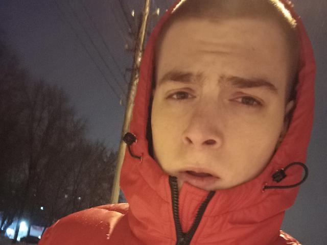 FreddieDillon with a confused expression, wearing a red hooded jacket, has a fair skin tone and brown hair. Nighttime, streetlights in background, slightly wet face.