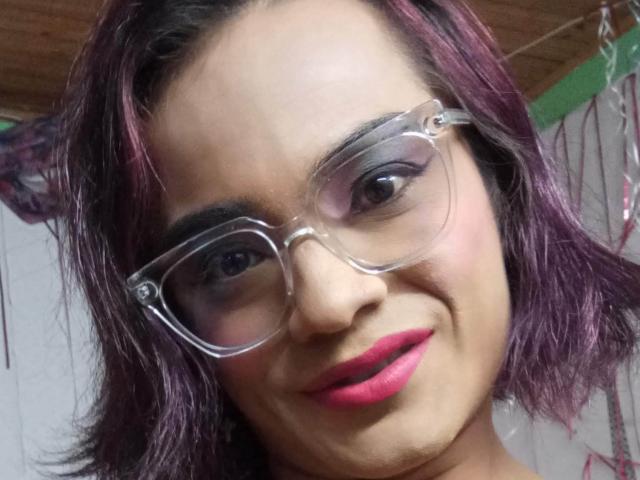 ChicoFemboy ha il rossetto rosa, gli occhiali trasparenti e i capelli tinti di viola, sorride con gli occhi marroni e un leggero sorriso. Il seno non è visibile, ma il viso è al centro dell'immagine..