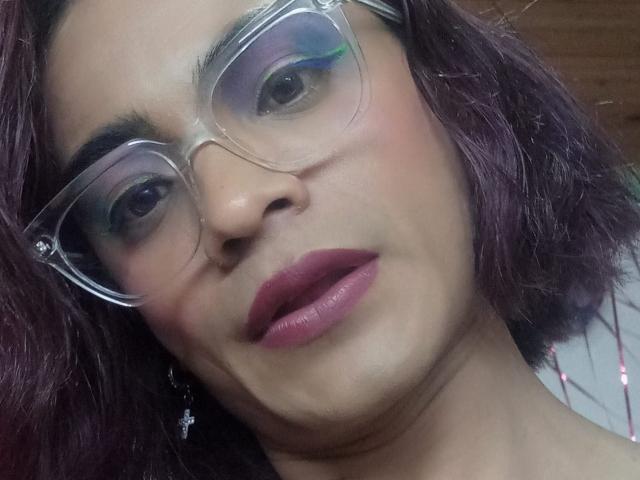 ChicoFemboy tem o cabelo tingido de roxo, usa óculos de grau e batom rosa escuro. Eles estão olhando para a câmera com um leve sorriso, mostrando um rosto fofo e feminino..