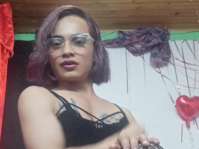 ChicoFemboy con cabello morado, gafas y un sujetador de encaje negro, mostrando los pezones, de pie en una habitación de madera, mirando a la cámara con una ligera sonrisa..