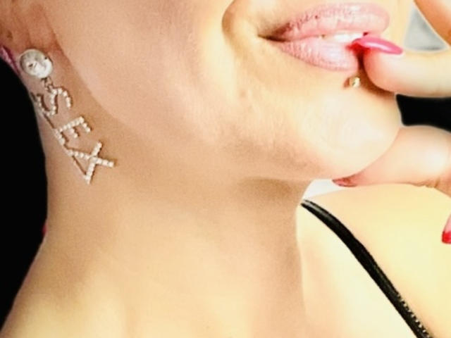 Le visage de KiwiJuicy, avec du rouge à lèvres rose, mordant une pointe d'ongle rouge, portant une boucle d'oreille "XOXO" incrustée de diamants à son oreille gauche, et vêtue d'un haut noir à bretelles..