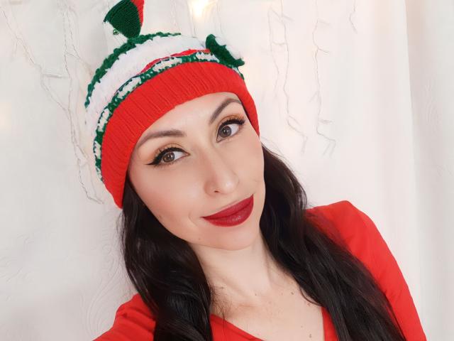 AliceRossi with red lipstick, green and white knit elf hat, and red top, has dark wavy hair, light skin, and brown eyes, smiling softly. Her tits are not visible.