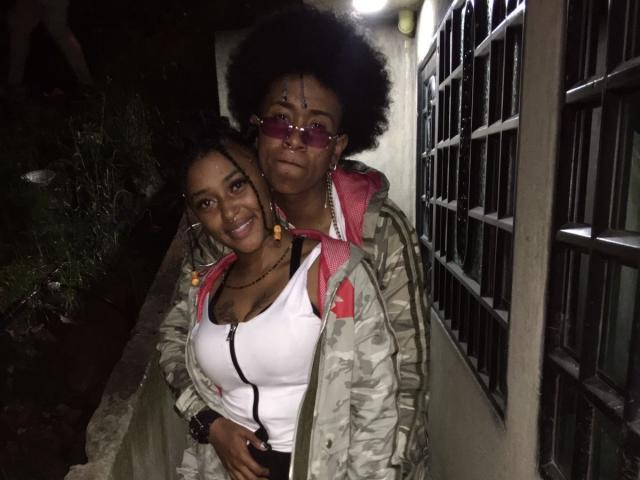 RileyXJohn, an older woman with big afro and glasses, hugs a younger woman with big tits in a white tank top and camouflage jacket, standing outside at night.