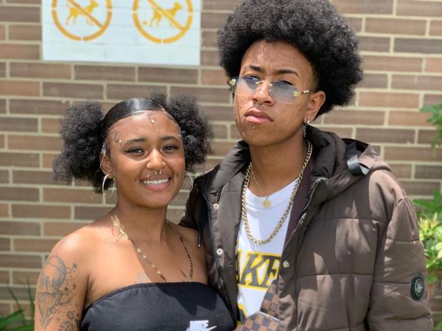 RileyXJohn, com um afro grande, óculos e piercing no nariz, vestindo uma jaqueta marrom, está ao lado de RileyXJohn, com cabelo preto volumoso, brincos de argola e uma blusa preta com alças..