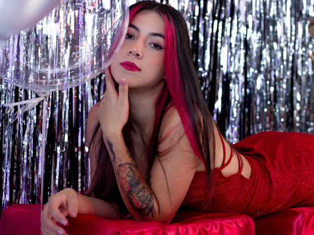 JuliaGarciax with pink and black hair, red lipstick, nose ring, and tattoo on her left arm, lying on red fabric, finger on her chin, looking at camera.