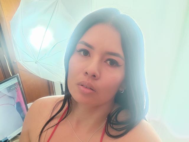AshleyKatelyn, com cabelos pretos longos, lábios carnudos e delineador, vestindo uma blusa vermelha, usa um colar de prata, e um guarda-chuva branco está ao fundo..