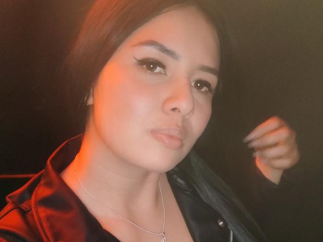 AshleyKatelyn with medium skin, black hair, subtle makeup, winged eyeliner, nose ring, black jacket, and necklace, looking at camera with a slight pout. Orange light on her face.