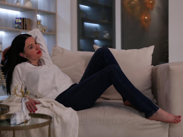 HottAlina, with black hair and red lipstick, lounges on a beige sofa in a white sweater and dark jeans, legs crossed, arms behind head, barefoot, looking relaxed.