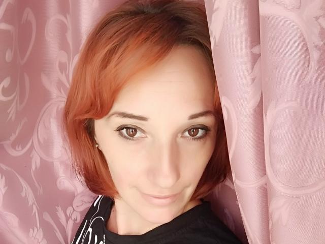 MissieLorrie, com cabelo ruivo, olhos castanhos e um leve sorriso, vestindo uma camisa preta, está em frente a cortinas rosa. Ela está olhando diretamente para a câmera..