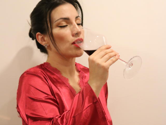 Liliysweet, avec ses cheveux noirs attachés en chignon, portant une robe rouge en satin brillante, sirotant du vin rouge dans un verre transparent, les yeux fermés, rouge à lèvres rouge, petit anneau de nez et boucles d'oreilles..