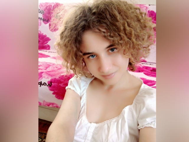 SelenaXCute, con cabello rubio rizado, ojos azules y una blusa blanca, mostrando una ligera sonrisa, con senos pequeños visibles y un rostro adorable, de pie frente a un fondo floral rosa..