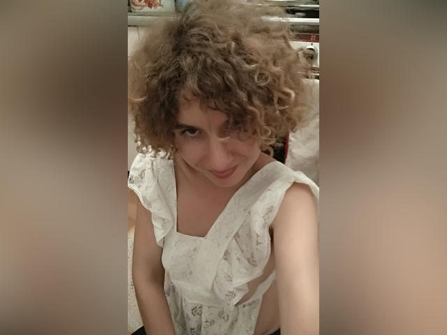 SelenaXCute, com cabelos castanhos cacheados, vestindo um vestido branco de renda, olhando para cima com um leve sorriso, mostrando seus pequenos seios e traseiro esguio em um selfie..
