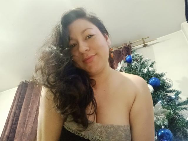 SakotoDika with wavy black hair, smiling, wearing a strapless grey lace top, showing her boobs, standing by a decorated Christmas tree with blue and white ornaments.