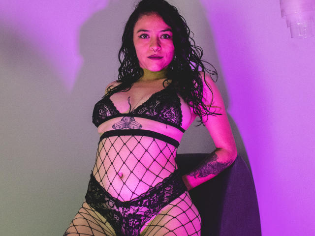 SamyBrook, con i capelli neri e umidi, indossa un reggiseno di pizzo nero, collant a rete e biancheria intima di pizzo abbinata, mostrando dei tatuaggi, con illuminazione viola e un'espressione sicura..