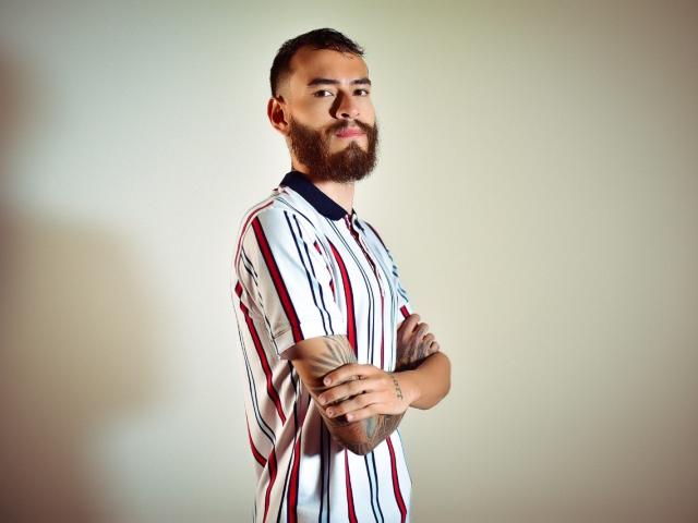 AndersX, con barba completa, brazos cruzados y una camisa a rayas, sosteniendo una pelota de béisbol, de pie frente a una pared lisa, con una expresión de confianza y una ligera sonrisa..