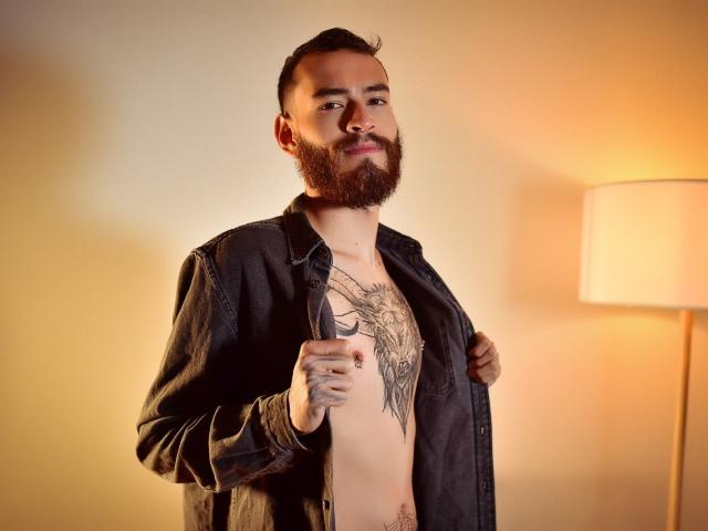 AndersX met een baard, shirt open, waardoor een grote tatoeage op zijn borst zichtbaar is, wijzend met zijn getatoeëerde hand, staand in de buurt van een lamp, lachend. Hij heeft geen shirt aan, maar draagt een zwarte denimjas..
