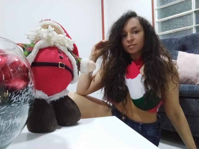 Angelonix with curly brown hair, wearing a red, white, and green crop top, sitting next to a stuffed Santa with a red hat and black belt on a white table.