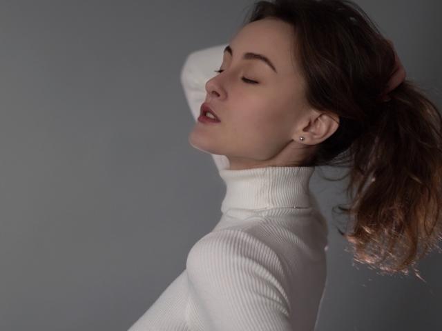 Alesya Drons, con los ojos cerrados, lleva un jersey de cuello alto blanco, tiene el pelo castaño recogido en una coleta, mostrando un pequeño piercing en la oreja, y tiene los labios rojos ligeramente entreabiertos..