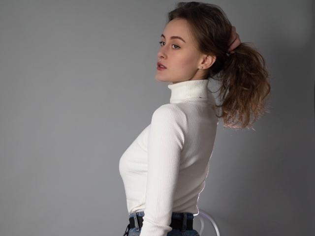 Alesya, com os lábios curvados, vestindo um gola alta branca, tem o rabo de cavalo castanho preso com um elástico rosa, mostrando seus pequenos seios e olhando por cima do ombro..