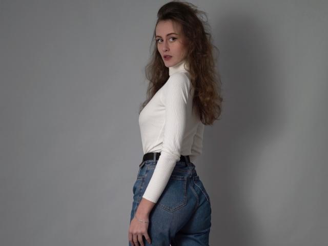 AlesyaDrons with wavy brown hair, white turtleneck, and blue jeans, standing sideways, showing her ass and looking over her shoulder with a slight pout.