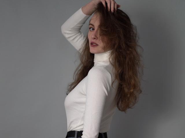 Alesya, com cabelo castanho ondulado, gola alta branca texturizada e calças pretas, olhando por cima do ombro, com a mão no cabelo, fundo cinza, luz suave. Seios visíveis sob a gola alta..