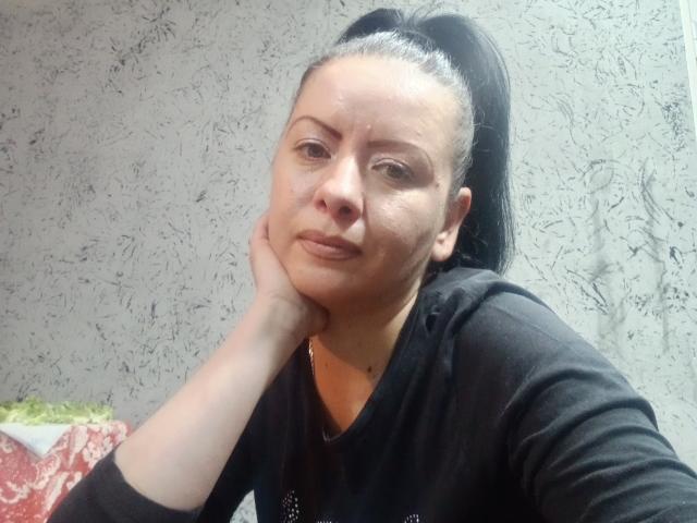 VioletaSwitch, con cabello rojo rizado, gafas azules y lápiz labial morado oscuro, sosteniendo un cigarrillo cerca de su boca ligeramente abierta, vestida con una camisa roja, mirando hacia arriba..