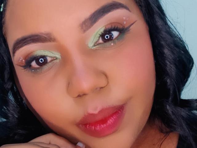 SarahSmity has green eyeshadow, black wavy hair, red lipstick, and a nose ring. Her eyebrows are thick, and she's looking up with a slight smile.