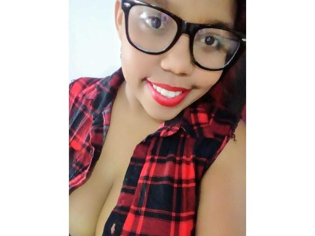SarahSmity, com seios grandes, vestindo uma camisa xadrez vermelha, óculos pretos e batom vermelho, sorrindo para a câmera, exibindo seu decote..