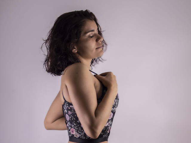 Bulma, con i capelli corti e mossi, scuri, indossa un abito nero con fiori, tiene le bretelle, vista laterale, leggero sorriso, occhi chiusi, mani sul petto, luce soffusa, sfondo chiaro..