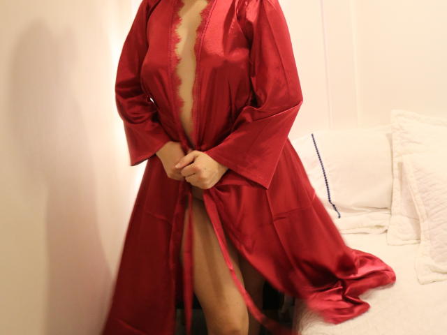 Liliysweet in a red satin robe, slightly open to reveal her bare tits and a peek of her pussy, standing by a bed with white pillows.