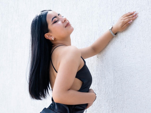 Olivia Monteiro mit schwarzen Haaren, geschlossenen Augen und einem Lächeln, trägt ein schwarzes Kleid, steht mit dem Rücken zur Kamera, die rechte Hand liegt auf einer weißen Wand, der linke Arm liegt über dem Bauch, ein silbernes Armband am Handgelenk..