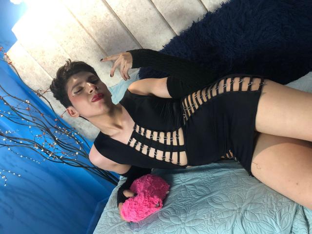 Saturni está deitada em uma cama, vestindo um vestido preto com recortes em listras, que revelam uma pequena parte das nádegas e um espaço entre as coxas, com batom vermelho e cabelo preto curto..
