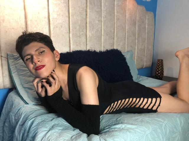 Saturni, com cabelo preto curto e batom vermelho, está deitada em uma cama usando um vestido preto com recortes, que revela seu corpo esguio e nádegas pequenas, com as mãos perto do rosto..