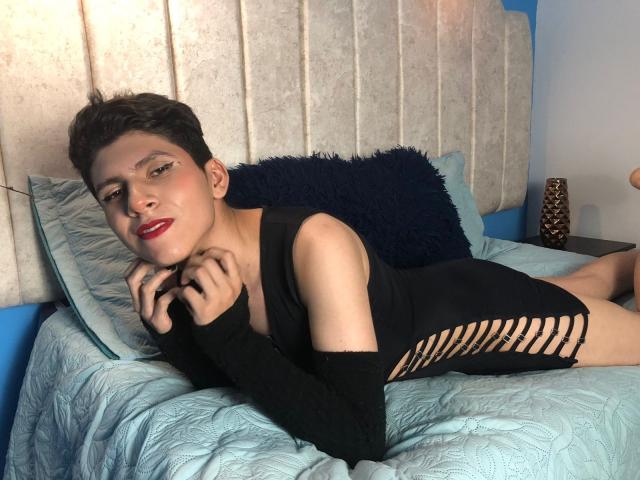 Saturni, com cabelo preto curto, batom vermelho, deitada na cama em um vestido preto com recortes laterais, mãos perto do rosto, cama acolchoada azul, travesseiro escuro, pele clara, fofa..