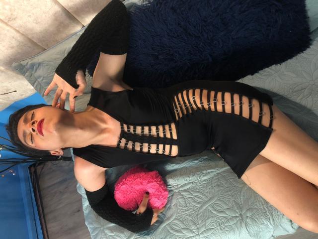 Saturni, vestida con un vestido negro con aberturas, está acostada en una cama con almohadas azules y rosas. Lleva mangas negras, lápiz labial rojo y sostiene un corazón rosa..