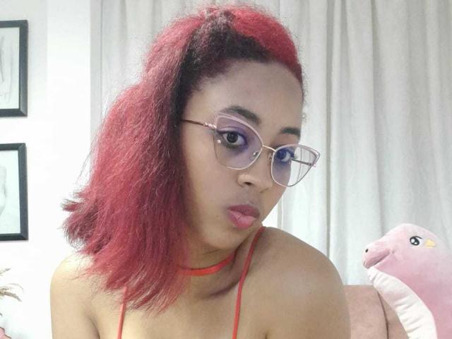 VioletaSteel, avec des cheveux roux, des lunettes et un collier rouge, les lèvres pincées, regardant la caméra, portant un haut rouge à encolure américaine, montrant ses seins. Un animal en peluche rose est visible sur la droite..