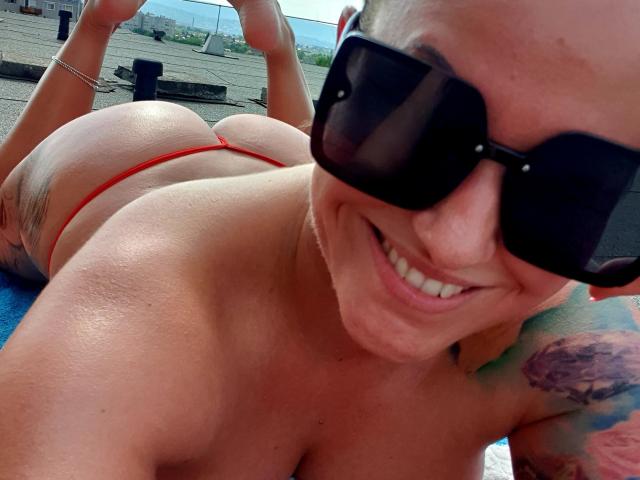 HotGirlVibe acostada boca abajo, con gafas de sol negras, tanga roja, sonriendo, mostrando su trasero, tatuaje en el brazo derecho, toalla azul, día soleado, piel bronceada, cabello rubio corto..