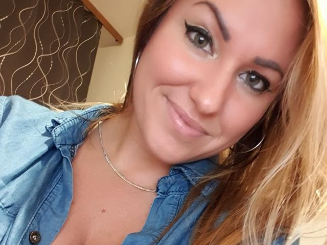 Una ragazza con un'aria seducente, con capelli castano chiaro, indossa una camicia blu, ha un sorriso carino, un trucco leggero, eyeliner, mostra un po' di scollatura e porta una sottile collana..