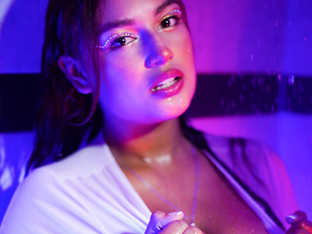 MallorySandler has wet brown hair, purple and blue neon lighting on her face, shiny skin, white shirt, and a visible cleavage with a silver necklace. Her mouth is slightly open.