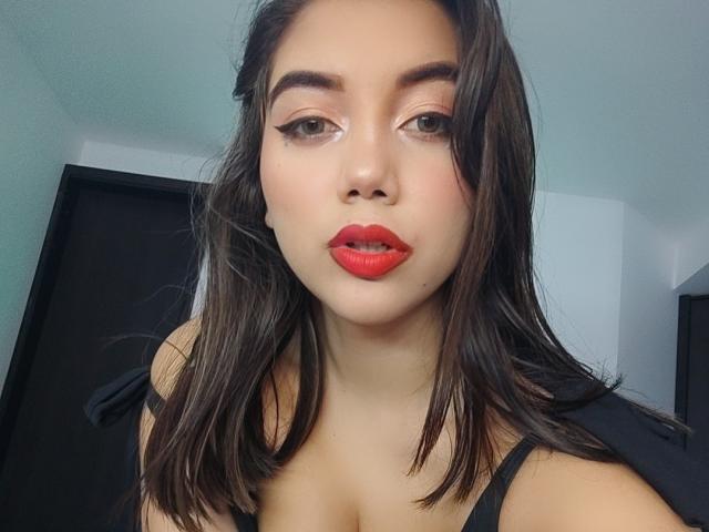 SquirtingCouple ha i capelli castano scuro, rossetto rosso e eyeliner nero, guarda direttamente la telecamera con un leggero sbuffo, indossando una maglietta nera che mostra il seno..