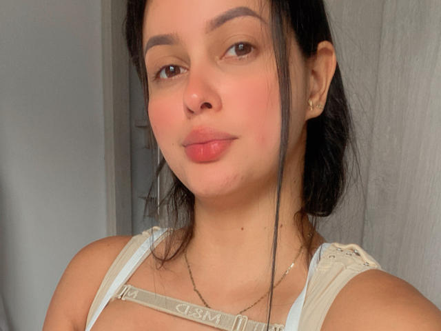 EmilyParkerr has light brown skin, dark eyebrows, and pink lips. She wears a white top with straps, a thin necklace, and has a small nose piercing. Her tits are visible.