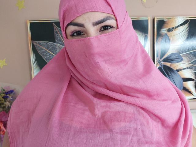 Sahily mit einem rosafarbenen Hijab, der ihr Gesicht bedeckt, wobei dunkle Augenbrauen und Augen sichtbar sind, trägt sie einen transparenten Stoff und blickt direkt in die Kamera, wobei weiches Licht ihre Gesichtszüge hervorhebt..