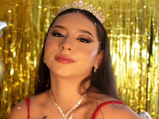 Mia Suarez, con el pelo largo y castaño, lleva una tiara brillante, un vestido rojo y un collar de diamantes. Tiene maquillaje con delineador de ojos oscuro y lápiz labial rosa, y se pueden ver tatuajes en su hombro derecho..