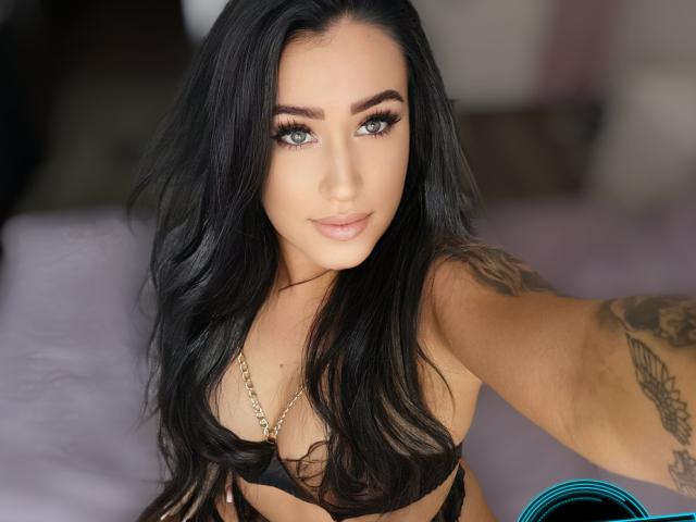 LovelyMichellex69, com cabelos pretos longos, olhos azuis e tatuagens, vestindo lingerie preta, olhando para a câmera com um leve sorriso, mostrando seus seios..