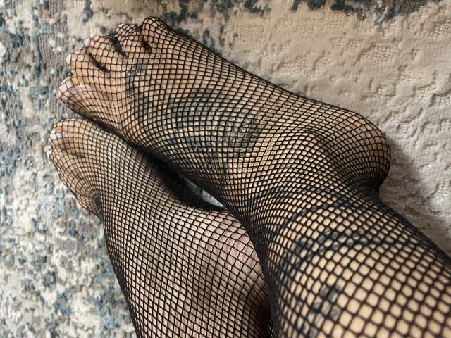 Misstyk's feet in black fishnet stockings, toes showing, on a textured rug. Her feet are crossed at the ankles, highlighting the net pattern over her skin.