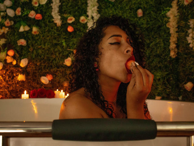 Scarlett, eine Frau mit lockigem, schwarzem Haar, dunkler Haut und rotem Lippenstift, lutscht an einem roten Lutscher und sitzt in einem dunklen, intimen Ambiente, wobei Kerzen im Hintergrund leuchten..